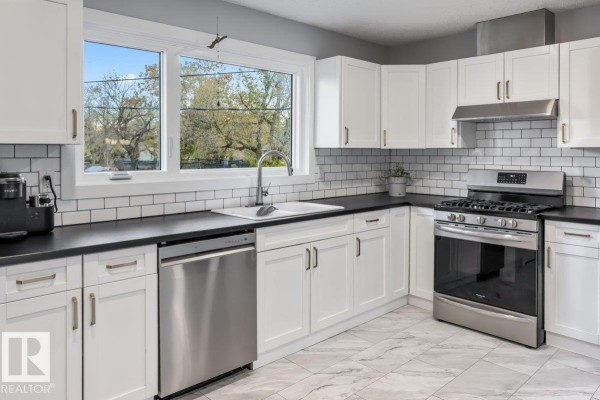 Modern white kitchen with stainless steel appliances, subway tile backsplash, and a large window. - 55 Gladstone Cr, St  Albert