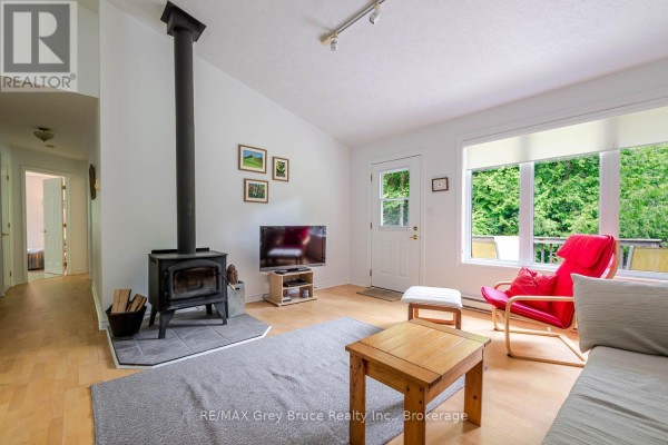 Living room with light wood floors, white walls, a black wood stove, and a large window overlooking greenery. - 550 Huron Road N, South Bruce Peninsula