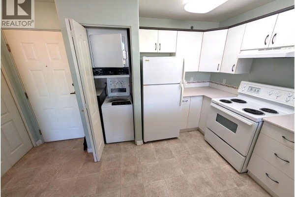Kitchen with white cabinets, white appliances, a stacked washer and dryer, and beige tiled flooring. - 554 Seymour Street Unit# 411, Kamloops