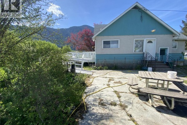 Concrete patio area with a wooden picnic table next to the house with overgrown bushes and trees. - 555 Carson Road, Grand Forks
