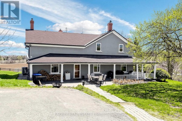 Rear view of a light blue farmhouse with a long wrap-around porch and chimneys. - 5626 3rd Line, New Tecumseth