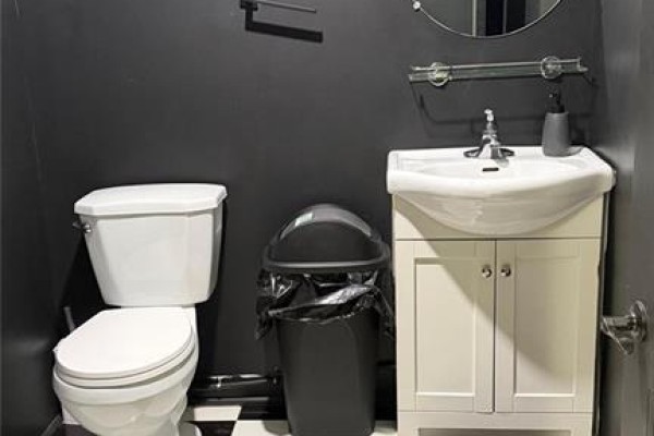 A small washroom with a white toilet, vanity sink, white cabinets, and a dark gray trash can. - 5657 Highway 7 Unit# 4, Vaughan