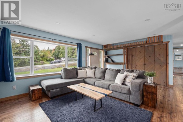 Living room with light blue walls, a grey sectional sofa, large window with view of trees and road, and wooden closet doors. - 57 Bayview Drive, Whites Lake