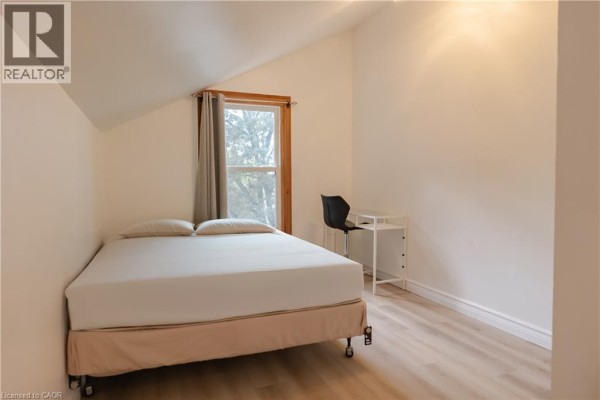 Bedroom with a bed, desk, and a window with grey curtains overlooking trees. - 57 Rupert Court Unit# Upper, Hamilton