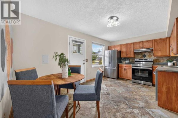 Dining area with a round wooden table and grey upholstered chairs, adjacent to a stainless steel kitchen. - 572 Regal Park Ne, Calgary