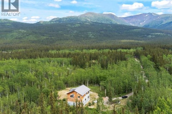 Wide aerial shot of a house in a forest clearing, with a gravel road and mountains. - 572 Robinson Subdivision, Whitehorse South