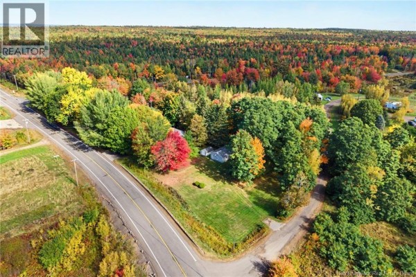 Aerial view of a property with a light gray house and metal roof, bordered by a paved road and dense forest. - 5724 Route 105, Mill Cove