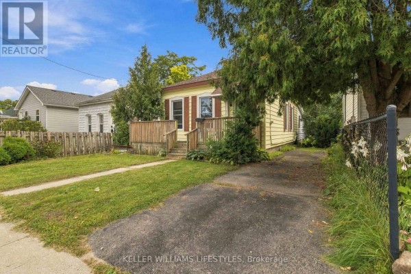 View from the side of the bungalow, showing the asphalt driveway, wooden fence, and mature trees. A white house is visible on the left. - 573 Hill Street, London East