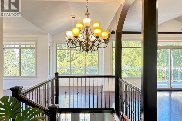 Interior staircase with dark wood railings and a chandelier above, overlooking large windows. - 5739 Bradbury Rd, Nanaimo