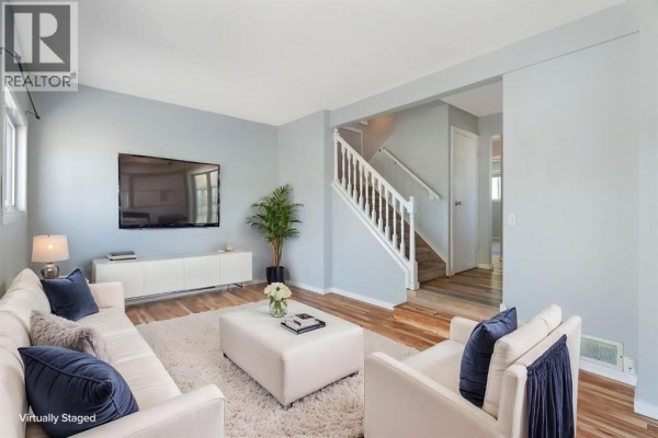 Living room with a white sofa, ottoman, wood floors, and staircase to the upper level. - 58, 5520 1 Avenue Se, Calgary