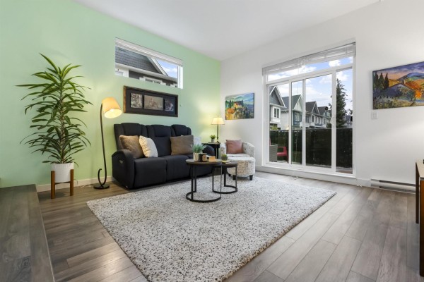 Living room with light wood floors, dark grey sofa, and sliding glass doors overlooking townhouses. - 58 16678 25 Avenue, Surrey