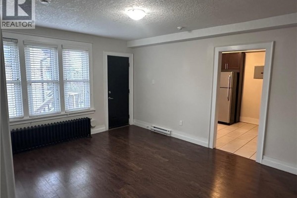 Living room with dark hardwood floors, white walls, and a black radiator under three windows. - 58-60 Madison Avenue N Unit# 203, Kitchener