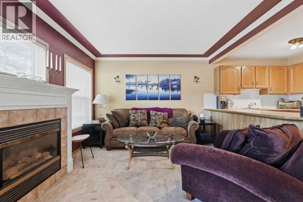 Living room with a gas fireplace, beige carpet, brown sofa, glass coffee table, and a small bar area. - 58 Country Hills Villas Nw, Calgary