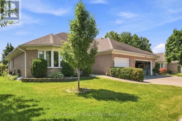 Side view of the brick bungalow with a green lawn, mature tree, and paved driveway. - 58 Ilderbrook Circle, Middlesex Centre