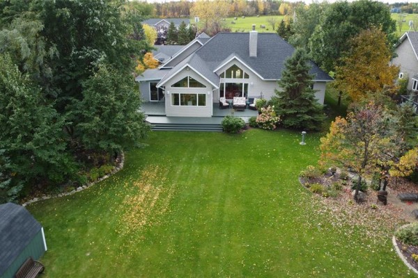 Aerial view of a backyard with a large green lawn, mature trees, and a light grey deck with patio furniture. - 58 Royal Road, Gimli