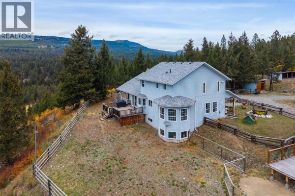 Aerial view of a light blue house with a large wooden deck and surrounding forested hills. - 5809 Beech Road, Merritt