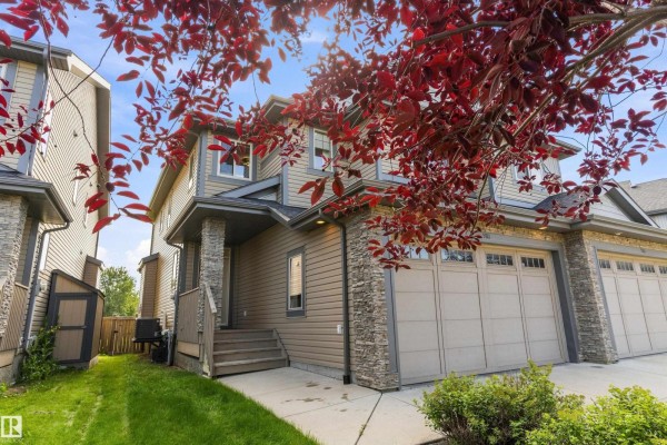 Side view of the duplex exterior with a grass lawn, red-leafed tree, and stone pillars. - 5811 Mullen Pl Nw, Edmonton