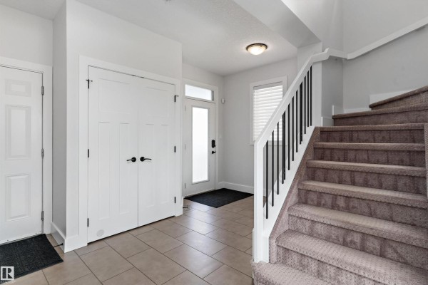 Foyer with tiled floor, white walls, white doors, and a carpeted staircase with black railings. - 5811 Mullen Pl Nw, Edmonton