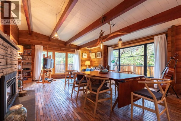 Living and dining area with wood walls and beamed ceiling, featuring a fireplace, large windows, and furniture. - 5817 Eagle Island, West Vancouver