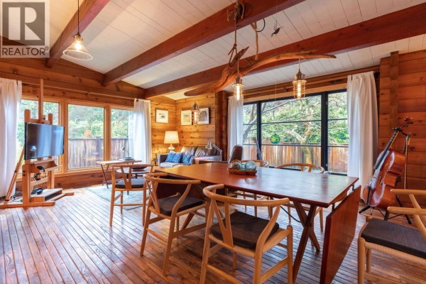 Interior shot of a wood-paneled dining and living area with a large table, fireplace, and views of trees. - 5817 Eagle Island, West Vancouver