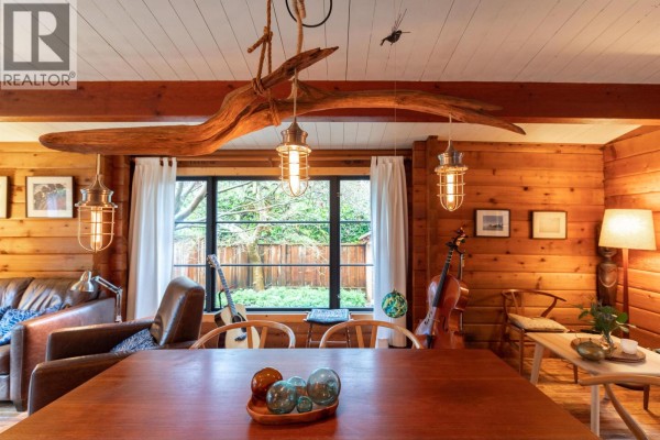 Close-up of dining table and chandelier in a wood-paneled room with large windows showing greenery outside. - 5817 Eagle Island, West Vancouver