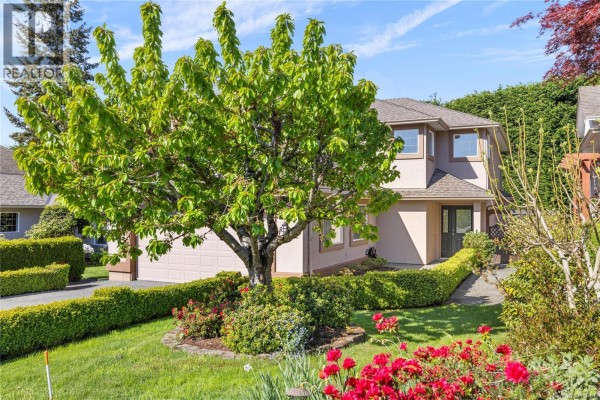 Front exterior of a house with a large green tree and red flowers in the foreground. - 5857 Quarry Cres, Nanaimo