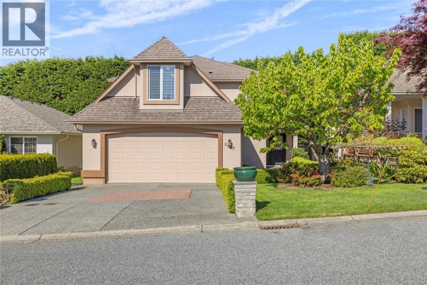 Front view of a house with a double garage door, beige stucco, and manicured landscaping. - 5857 Quarry Cres, Nanaimo