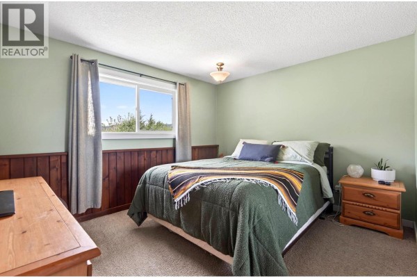 Bedroom with a green duvet, wood wainscoting, and a window with grey curtains. - 5890 Todd Road, Kamloops
