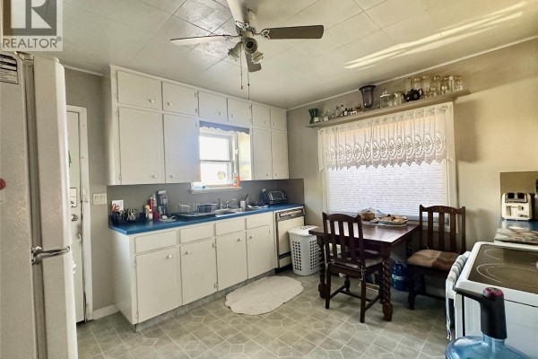Kitchen with white cabinets, blue countertops, small dining table, and a window with lace curtains. - 5952 Okanagan Street, Oliver