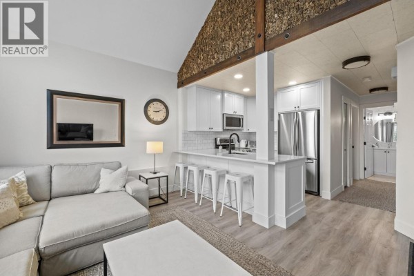 Living area with a sectional sofa facing a white kitchen island with stools and stainless steel appliances. - 5977 Snow Pines Crescent Unit# 116c, Big White