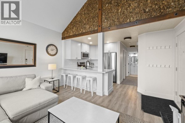 Living area and kitchen with white cabinets, island with stools, sectional sofa, and textured wall paneling. - 5977 Snow Pines Crescent Unit# 116c, Big White