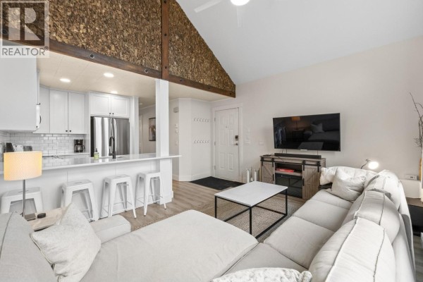 Living room with a sectional sofa, coffee table, television, and a glimpse of the kitchen area. - 5977 Snow Pines Crescent Unit# 116c, Big White