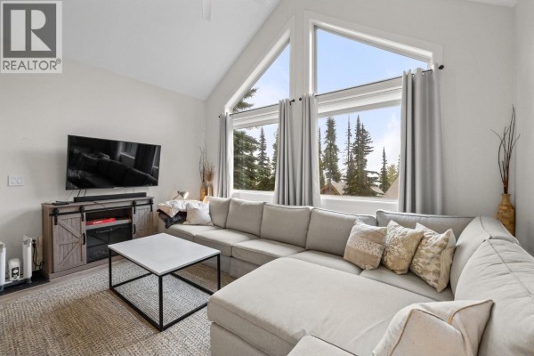 Living room with a large sectional sofa, coffee table, television, and expansive windows overlooking trees. - 5977 Snow Pines Crescent Unit# 116c, Big White