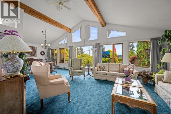 Living room with vaulted ceiling, exposed beams, blue carpet, and large windows with ocean views. - 5989 Garvin Rd, Union Bay