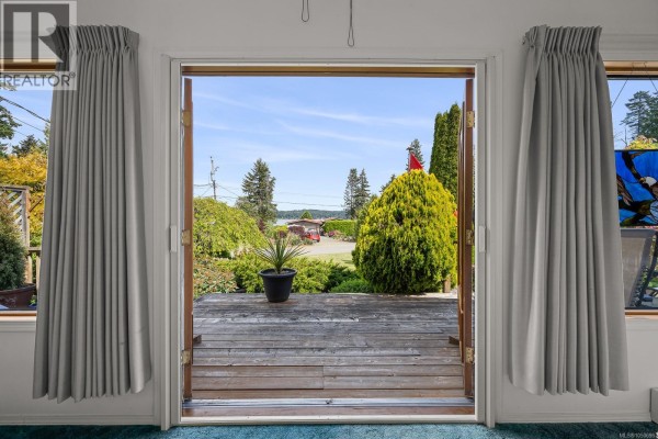 View from inside looking out double doors to a wooden deck and a garden with a distant water view. - 5989 Garvin Rd, Union Bay