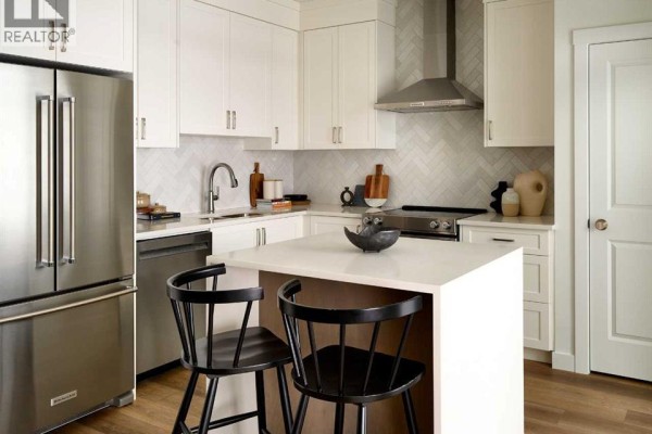 Kitchen island with two black stools, white countertops, and a view of the stainless steel refrigerator. - 6, 20830 Main Street Se, Calgary