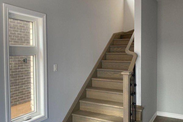 Wooden staircase with metal railings leading up, next to a tall narrow window with a brick exterior view. - 6 - 5672 Dorchester Road, Niagara Falls