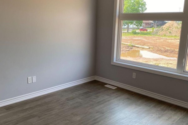 Empty room with grey walls, wood-look flooring, and a large window overlooking a muddy construction site. - 6 - 5672 Dorchester Road, Niagara Falls