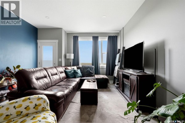 Living room with a large brown leather sectional sofa, television, and windows with dark curtains. - 6 3960 7th Avenue E, Regina