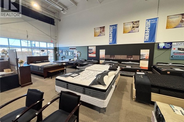 Wide view of mattress showroom with beds, furniture, and brand banners on the walls. - 6 805 Circle Drive E, Saskatoon