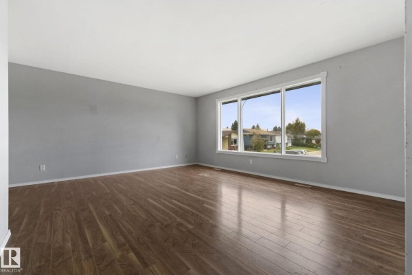 Empty living room with light grey walls, dark laminate flooring, and a large window with a view. - 6 Campbell Cres E, Stony Plain
