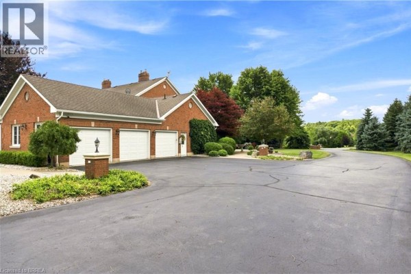 Side view of a brick house with a three-car garage, paved driveway, and surrounding trees. - 6 Hickory Hollow, Brant County