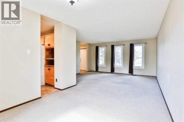 A large, bright living room with beige carpet, cream walls, and a row of four narrow windows with dark curtains. - 6 Temple Boulevard W, Lethbridge