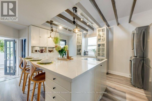 White kitchen with a large island, exposed wooden ceiling beams, and a stacked washer and dryer. - 6 Wheeler Avenue, Toronto