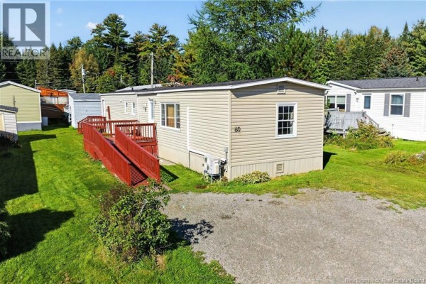 Street view of a beige mobile home with a large red wooden deck, surrounded by green grass, trees, and other homes. - 60 Midway, Moncton