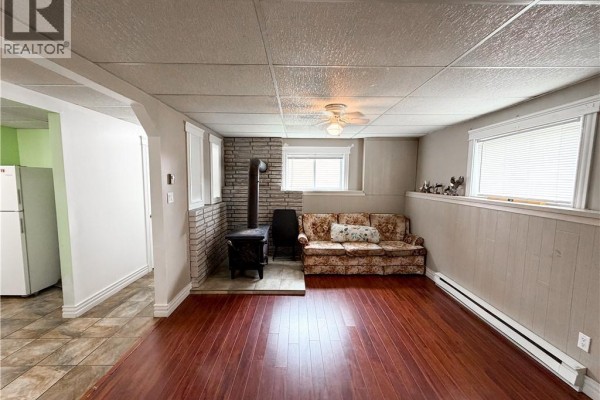 Living room with wood floors, a floral sofa, a wood stove on a stone hearth, and windows. - 60 Normandie Street, Petit Rocher