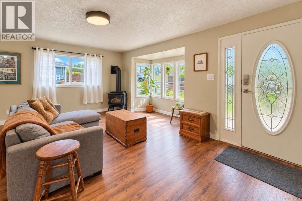 Living room with a wood stove, bay window, glass-panelled front door, and a grey sectional sofa. - 6008 Grandview Rd, Port Alberni