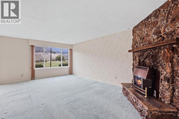 Living room with light blue carpet, large window with mountain view, and stone fireplace. - 6011 Dallas Drive, Kamloops