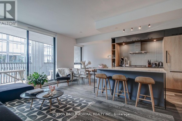 Living area with dark sectional sofa, patterned rug, armchair, and large window overlooking balcony. - 602 - 41 Dovercourt Road, Toronto