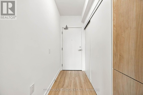 Interior hallway with light wood laminate flooring and a white apartment entrance door. - 602 - 7437 Kingston Road, Toronto